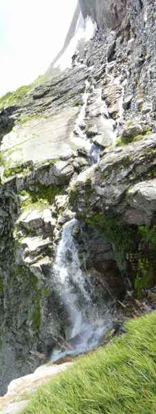 Cascade dangereuse psychédélique, prise en mode panorama diagonal la pente paraît beaucoup plus raide que la réalité (on peut s'en faire un idée en rétablissant la verticalité de la chute d'eau et des brins d'herbes), le danger ne réside pas tant dans la pente du chemin, mais plutôt en cas de course de vitesse à la descente avec petite distraction (le chemin fait un angle droit mal marqué et 4 mètres plus loin on est dans la cascade).